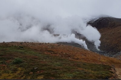 A geothermal landscape in Japan, showing steam rising from fumaroles on a rocky, barren mountainside. The foreground features patches of green and reddish vegetation, while thick clouds and mist partially obscure the background. A narrow trail winds through the terrain, leading toward the steaming vents, indicating volcanic activity typical of Japan's mountainous regions.