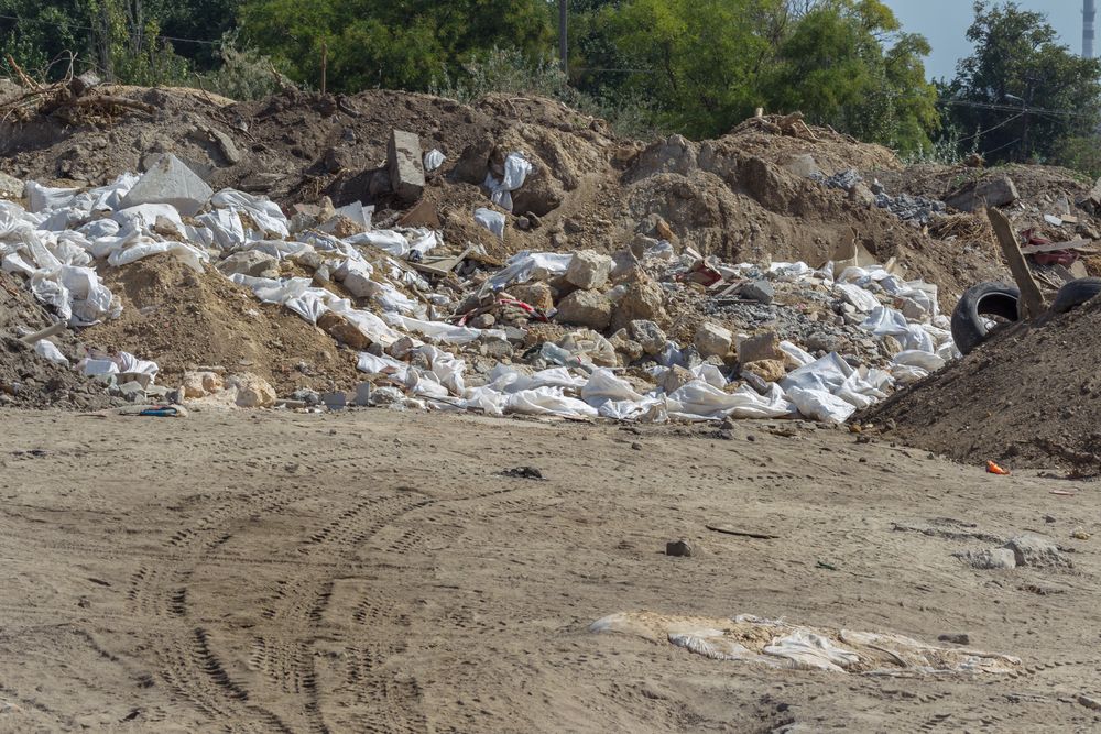 A pile of construction and demolition waste, including broken concrete, rocks, soil, white plastic bags, and discarded materials, dumped in an open area with tire tracks visible on the sandy ground.