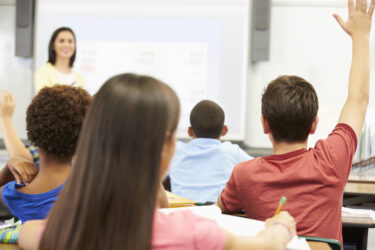 The image shows a classroom scene with a teacher standing at the front, smiling, and several students seated at desks facing her. Two students in the foreground have their hands raised, likely to answer or ask a question. The focus is on the students from behind, with the teacher slightly blurred in the background.